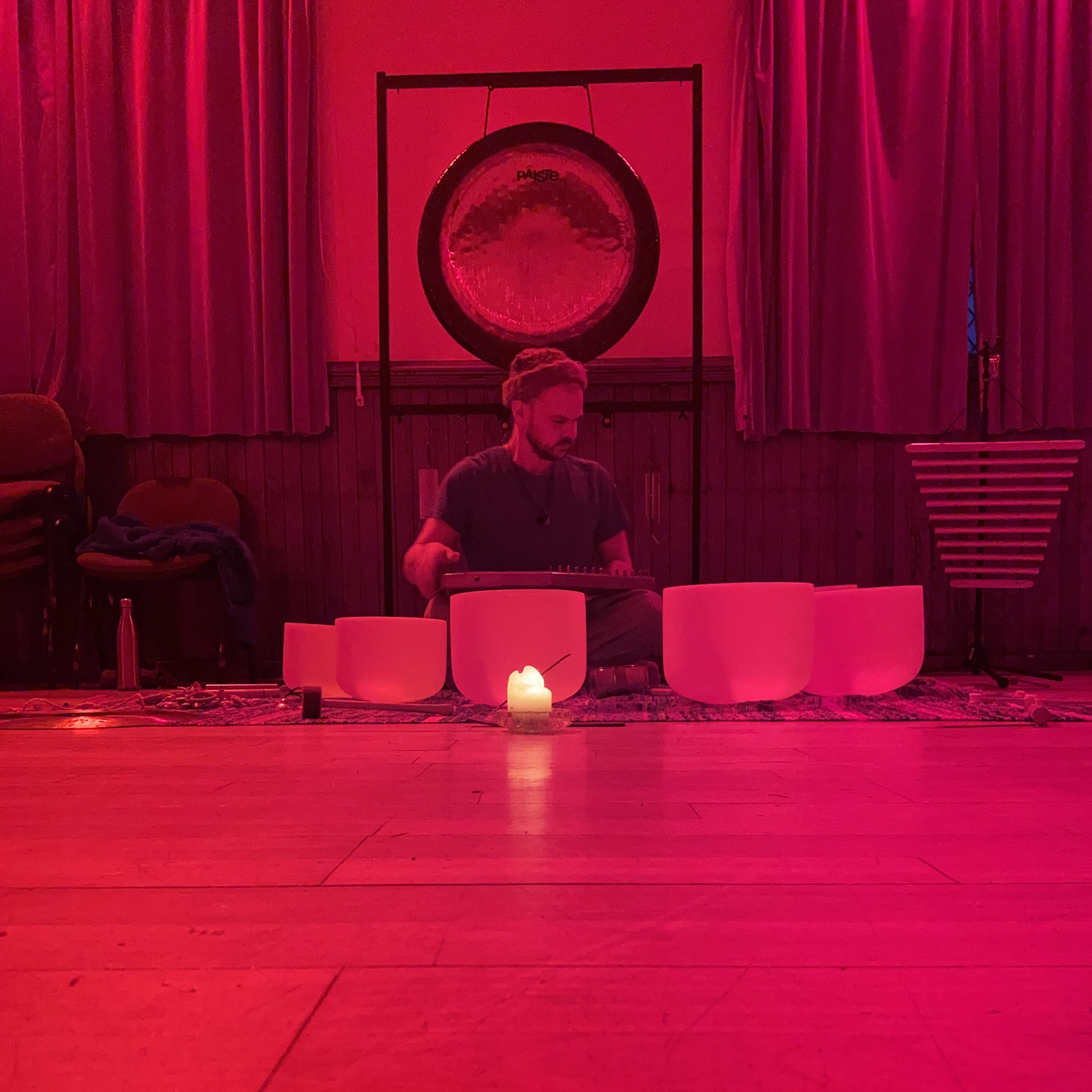 Room flooded in red light; Iain seated behind crystal bowls with a candle centered and a gong behind.