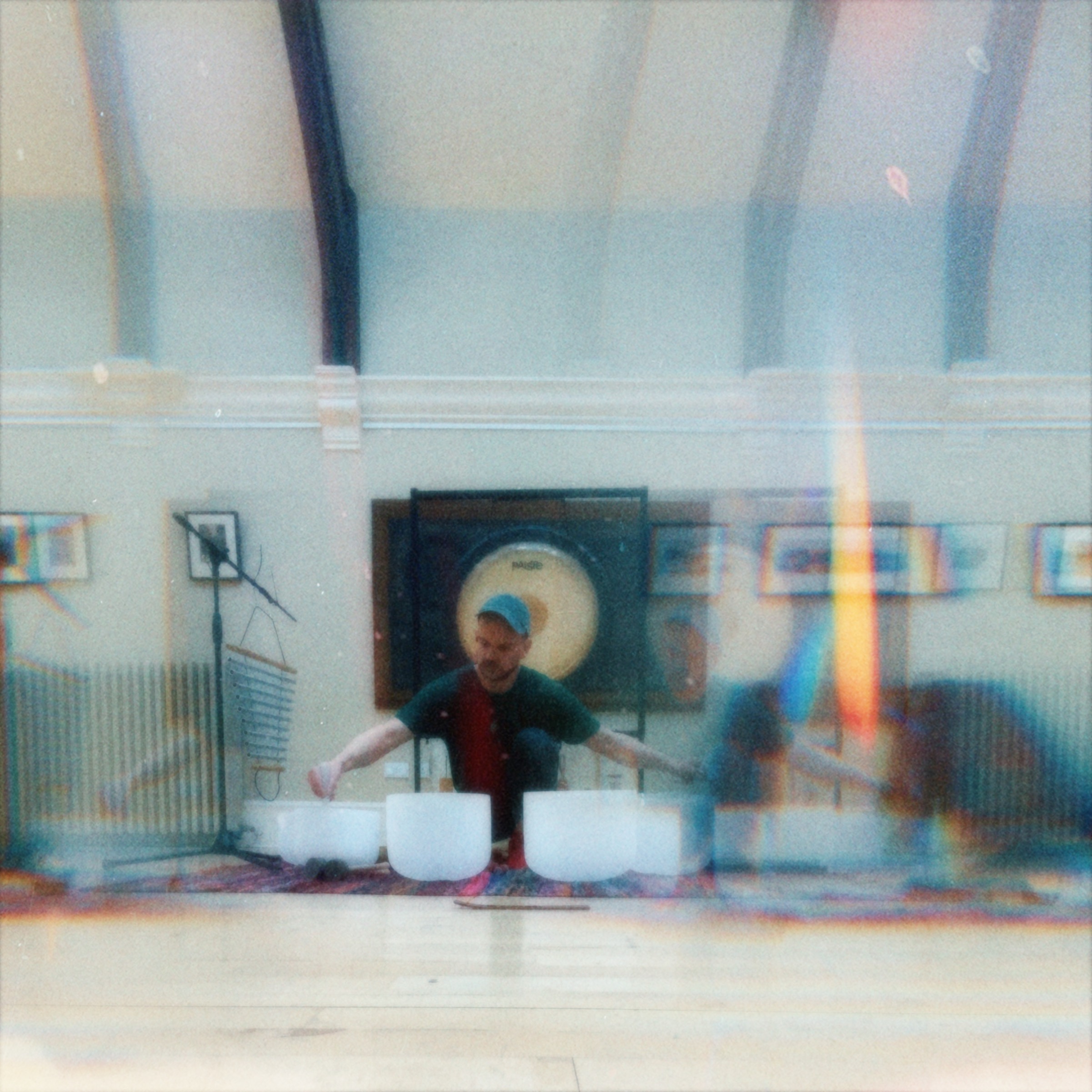 Iain performing with crystal singing bowls in a bright hall; large gong behind; dreamy light-leak effect.