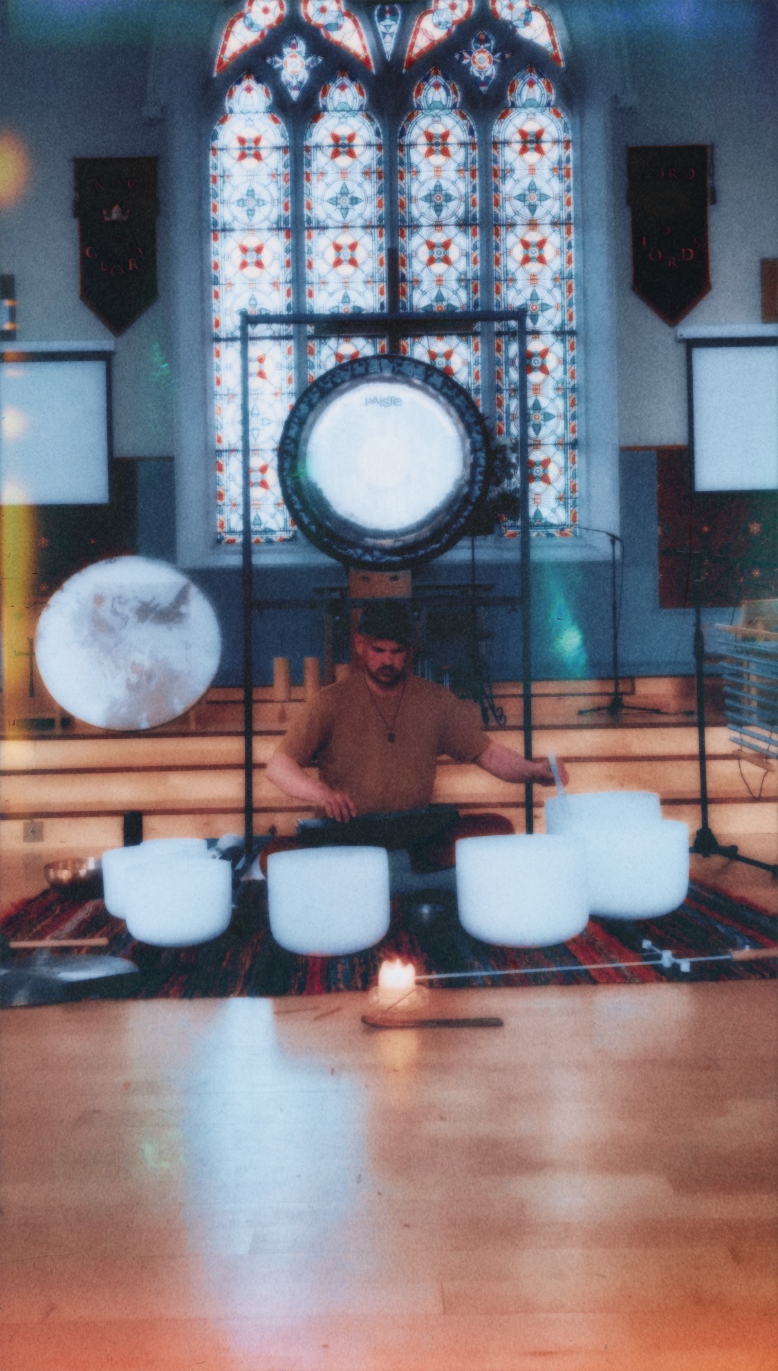 Iain performing surrounded by instruments under teal lighting; large gong and chimes behind.