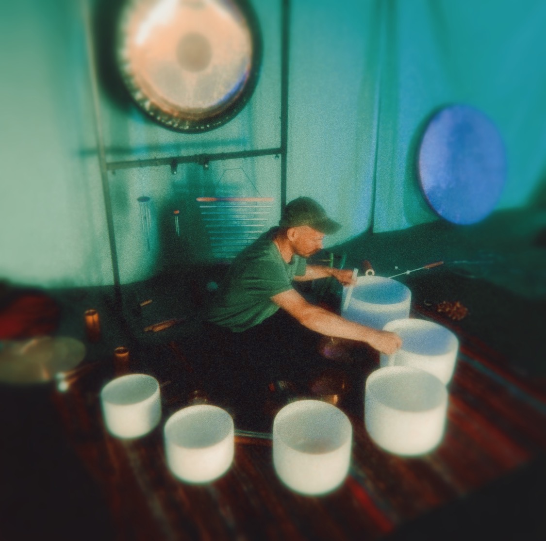 Side angle of Iain playing crystal bowls; teal drapes and a suspended gong above the setup.